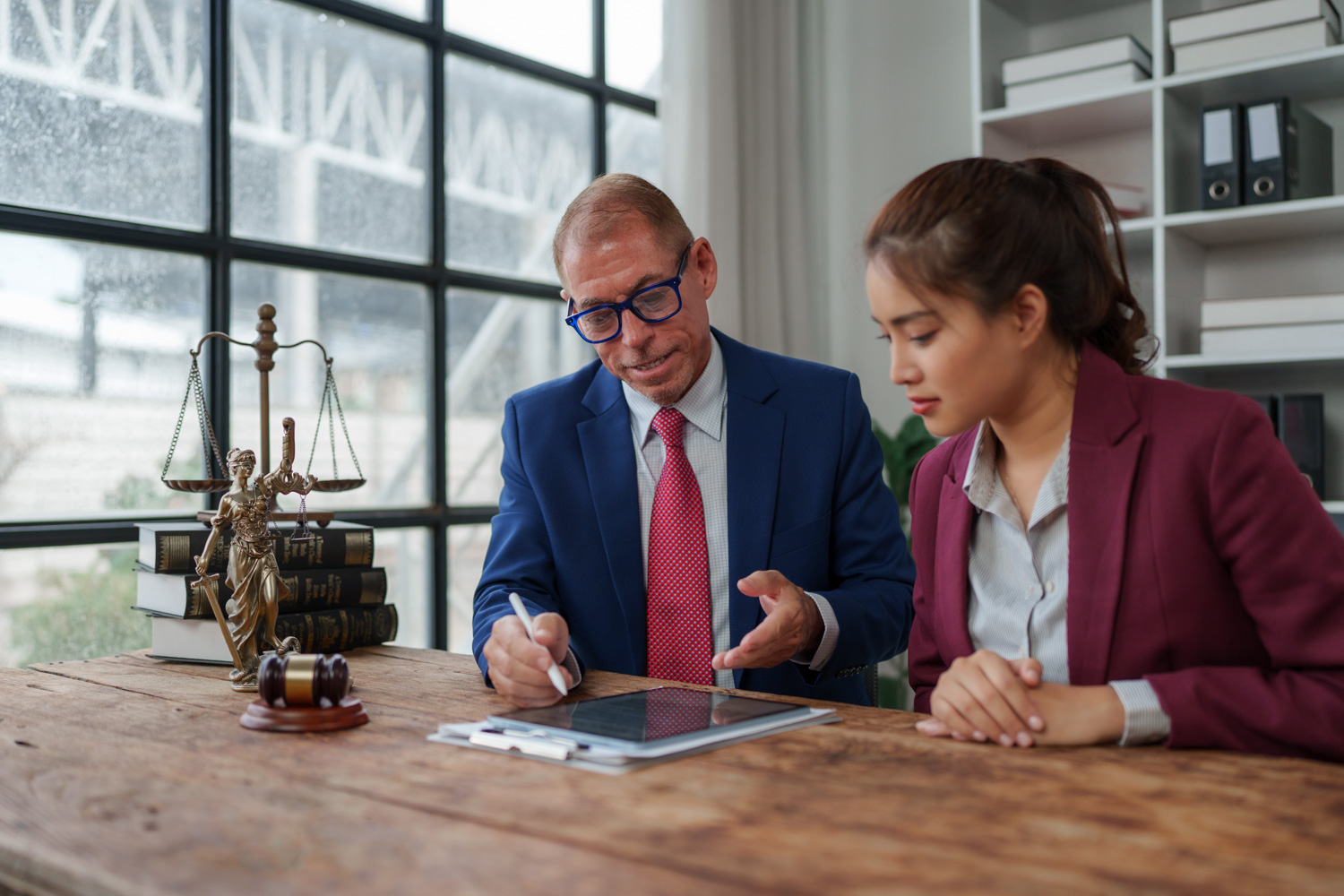 A law advisor and a student sit together at a desk reviewing information on a tablet in an office. A gavel, scales of justice, and law books on the desk emphasize the legal and academic advising setting.