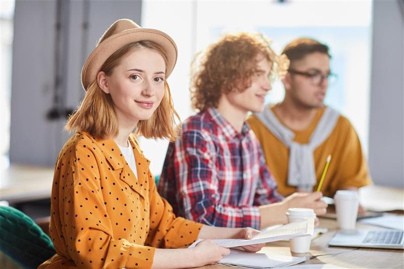Three students sitting at a table with their coffee, notes, and computers. A girl in yellow with a fedora is in focus, looking at the camera.