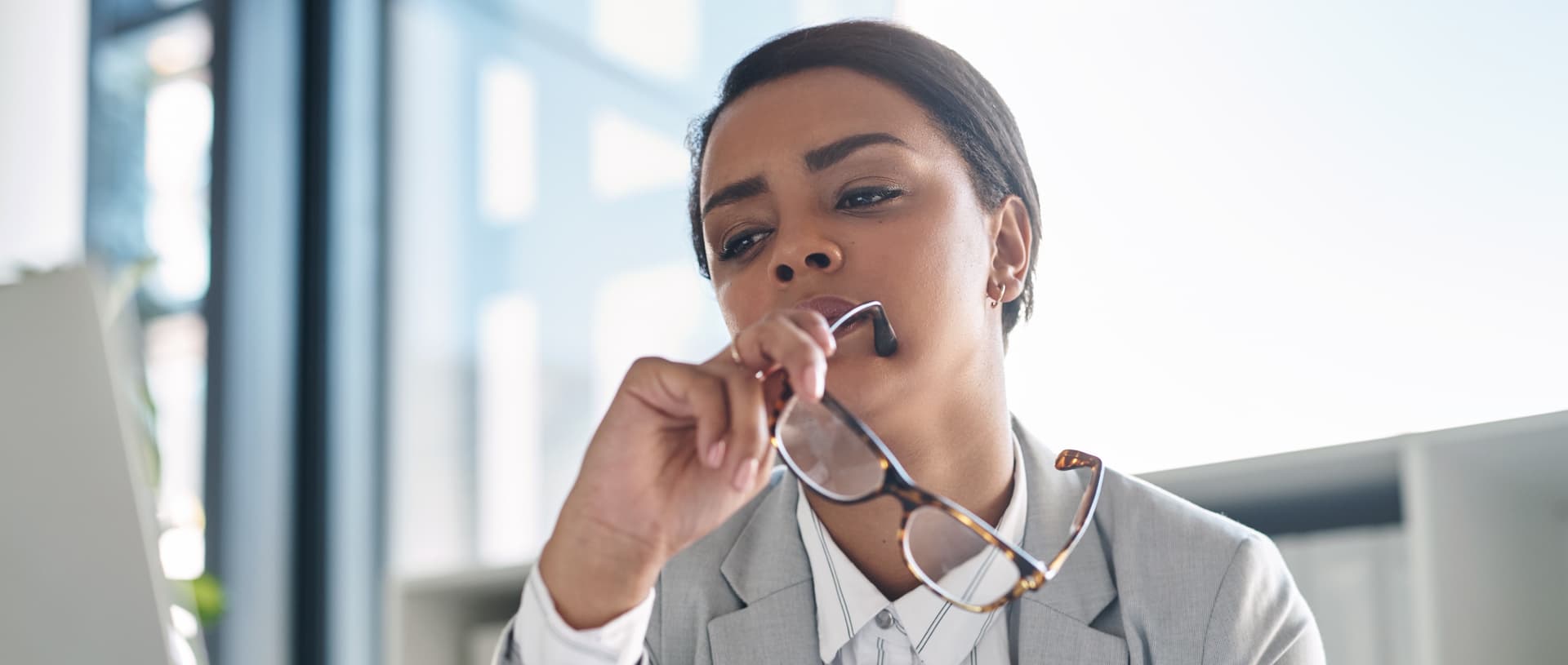 A professional woman sitting at her desk, thoughtfully considering her next steps while looking at a computer screen and holding her eyeglasses.