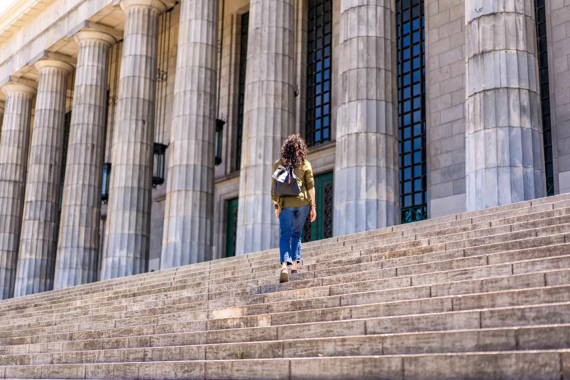 Individual with a bag pack climbing stairs to enter a building with large columns.