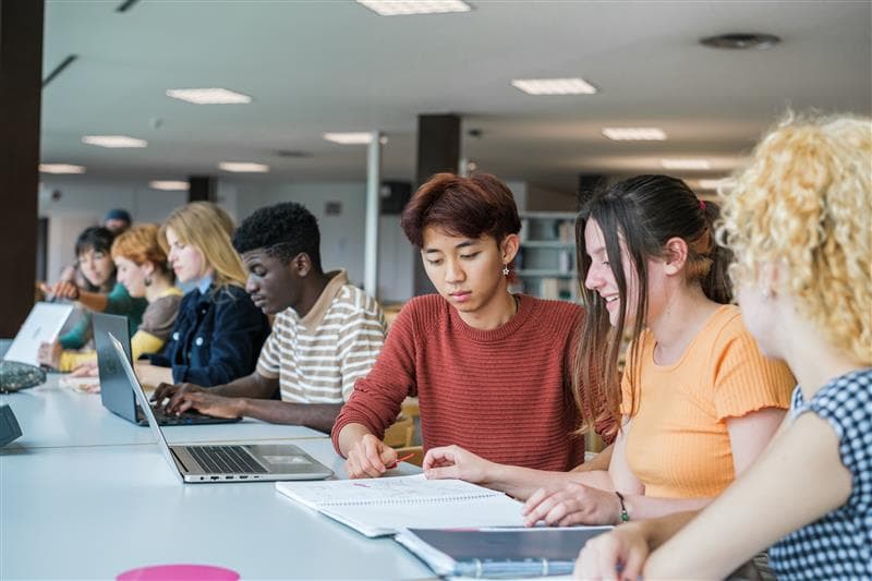 Three students sit at a desk with one student standing behind them — all looking at notes and sharing studying materials.